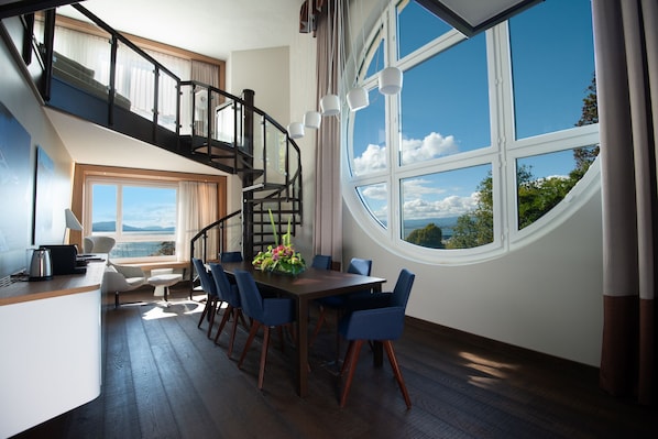 Un comedor con una mesa de madera y sillas azules, una escalera con barandillas negras, y ventanas amplias que ofrecen vistas al cielo y a los árboles.