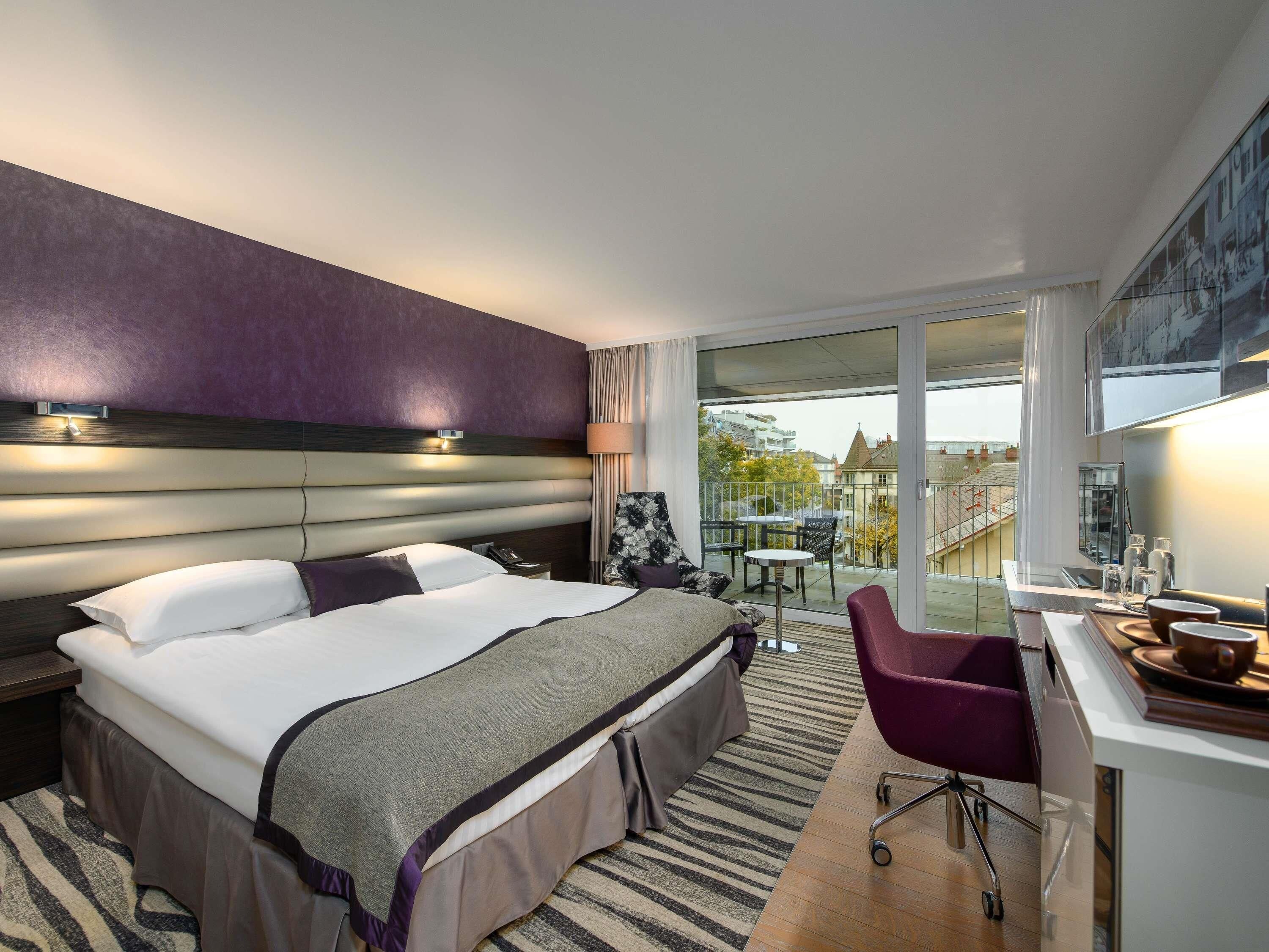 Habitación de hotel con una cama grande, un escritorio con silla, un balcón con mesa y sillas, y vista a la ciudad.
