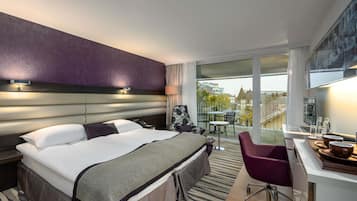 Habitación de hotel con una cama grande, un escritorio con silla, un balcón con mesa y sillas, y vista a la ciudad.
