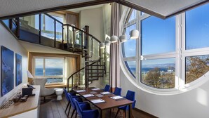 Habitación de hotel moderna con una amplia mesa de comedor, sillas azules, una escalera y grandes ventanales con vistas al mar.