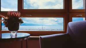 Habitación con un amplio ventanal con vistas a un puerto deportivo, una planta en maceta sobre una mesa y una silla orientada hacia la ventana.