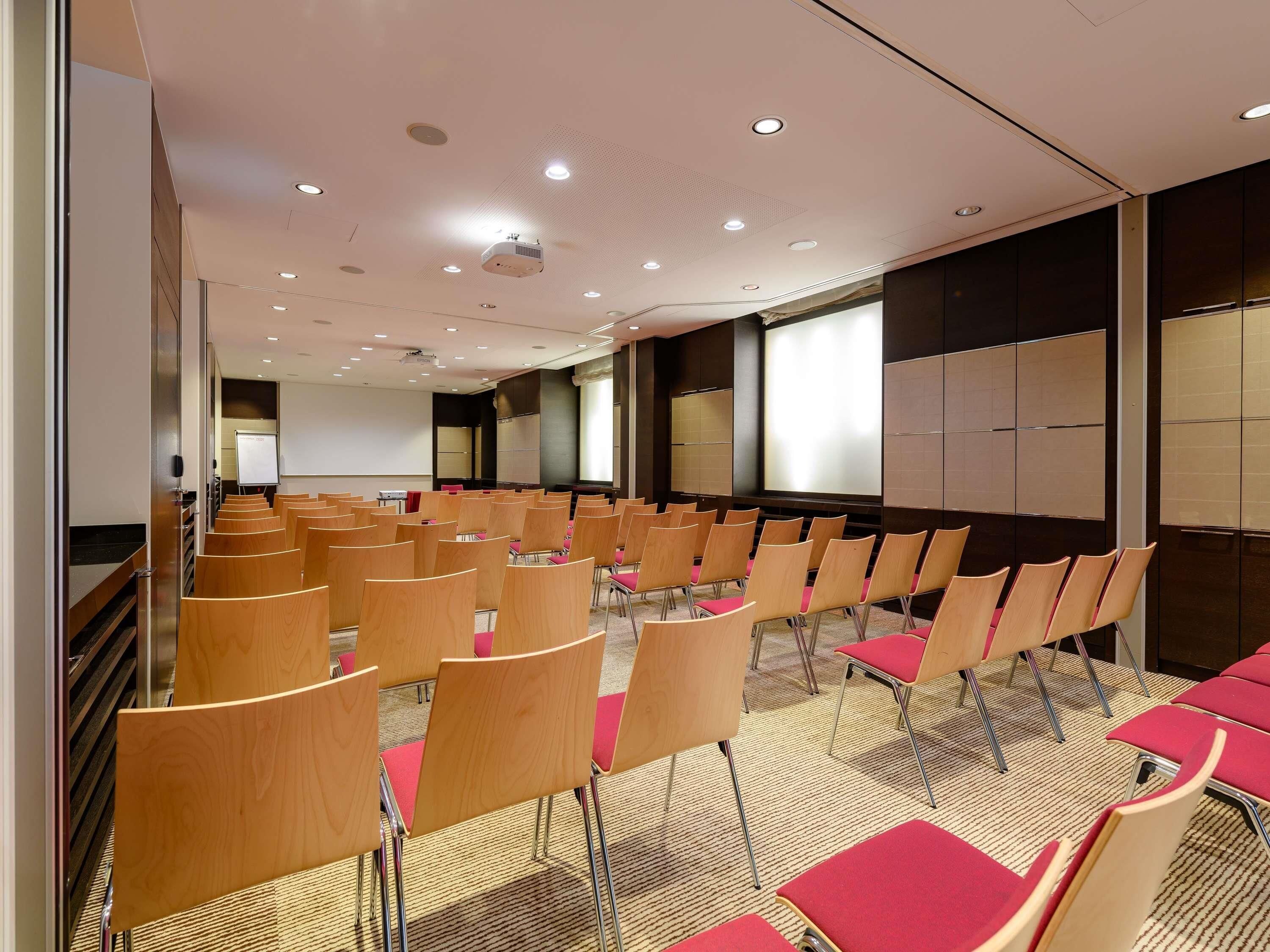 Une salle de conférence avec des rangées de chaises en bois et des coussins rouges, un projecteur et un grand écran.