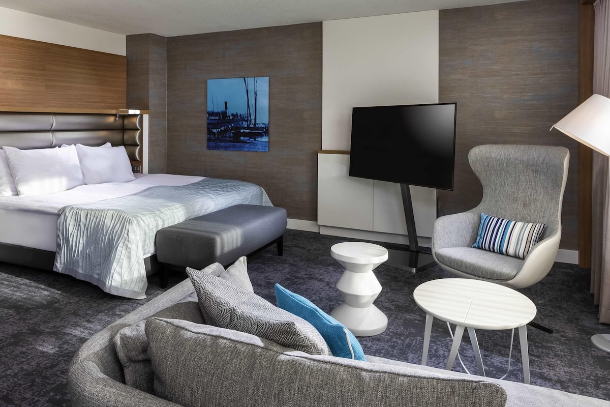 Une chambre d’hôtel moderne avec un grand lit, un téléviseur à écran plat, un fauteuil gris, une petite table ronde et un canapé agrémenté de coussins à motifs.