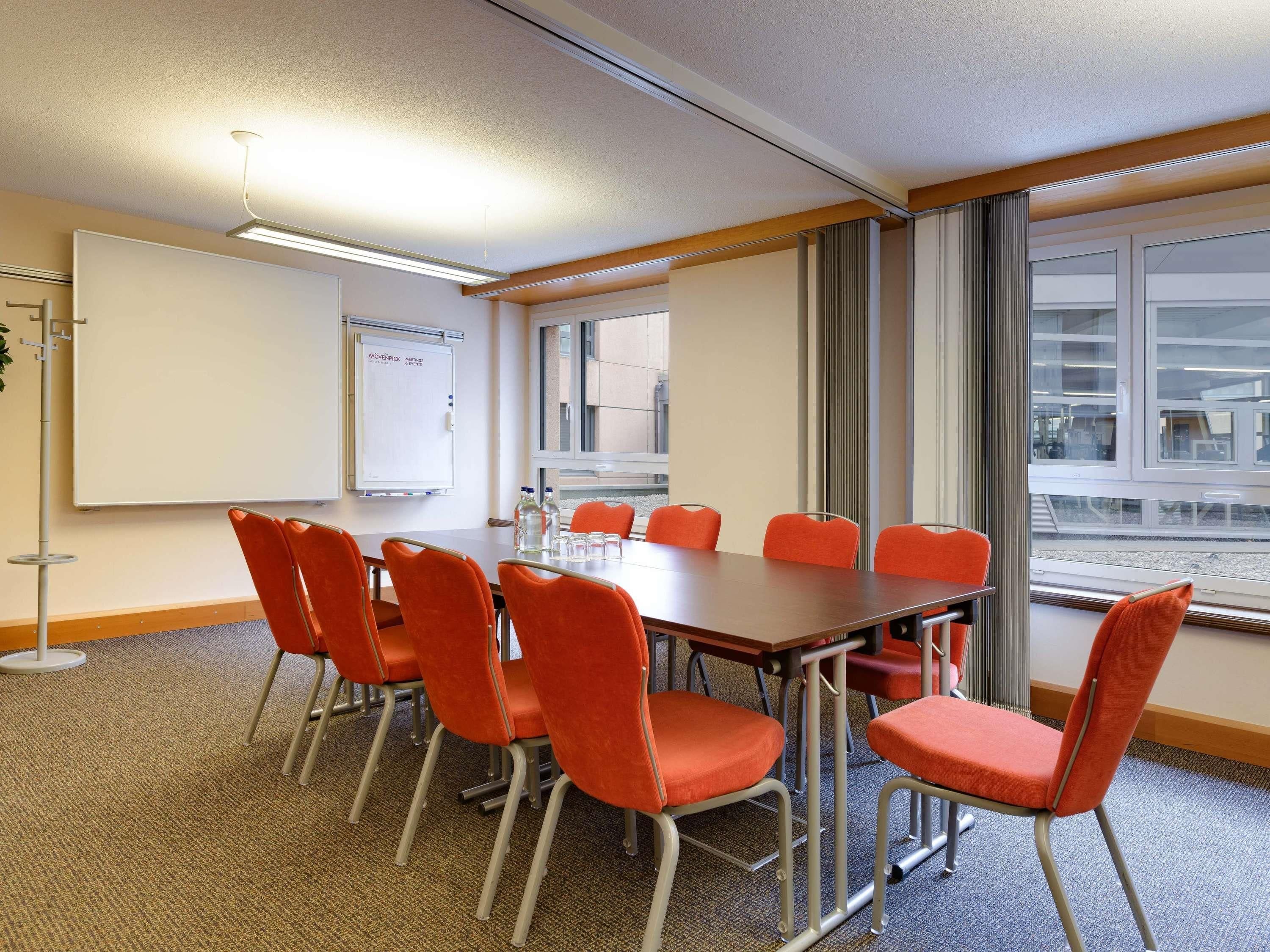 Un salón de conferencias con una larga mesa, seis sillas anaranjadas, una pizarra y grandes ventanales con vistas a edificios.