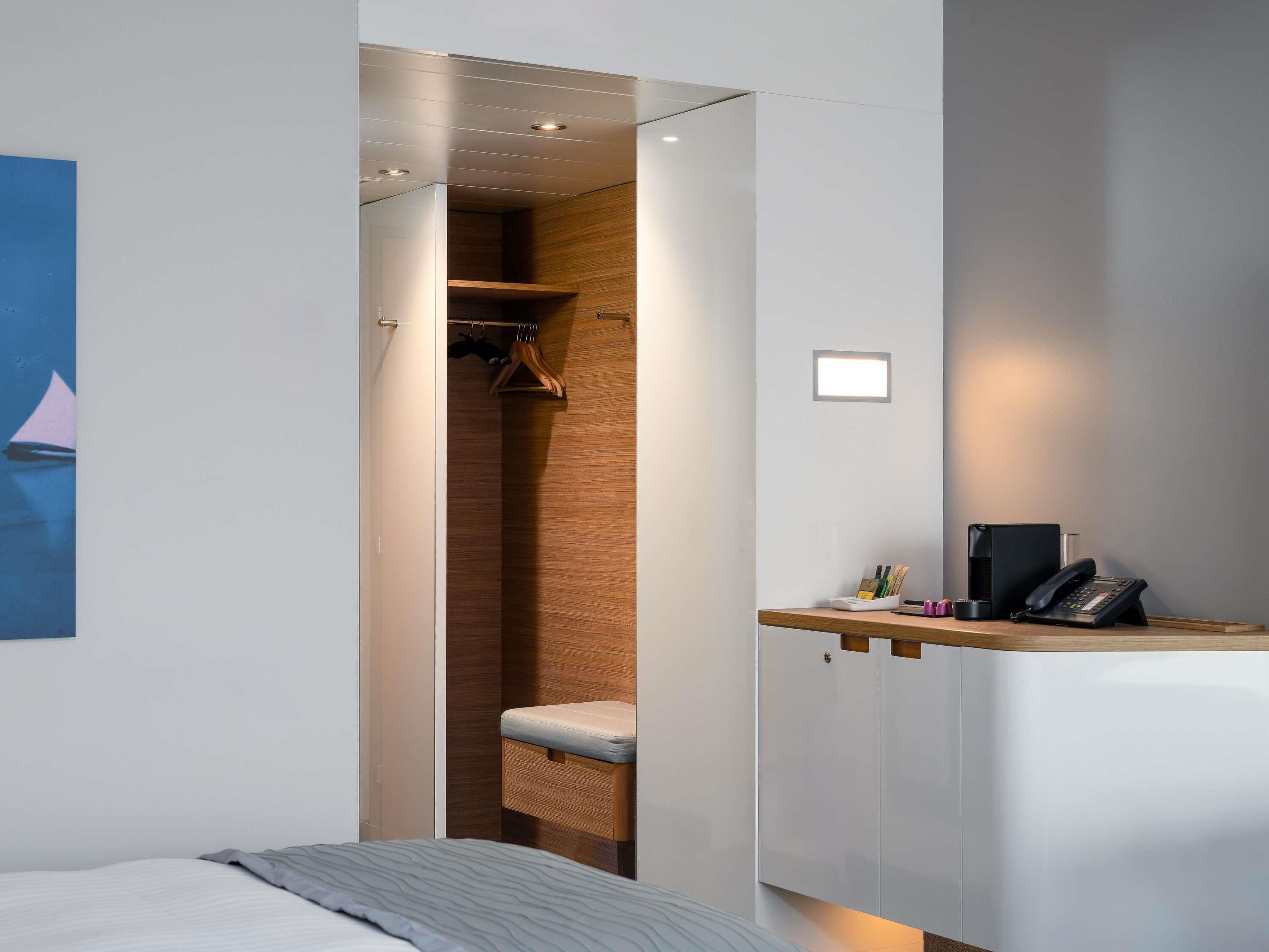 Ein kleines Hotelzimmer mit einem Bett, einem Schreibtisch mit Telefon und einem Holzschrank mit Ablage.