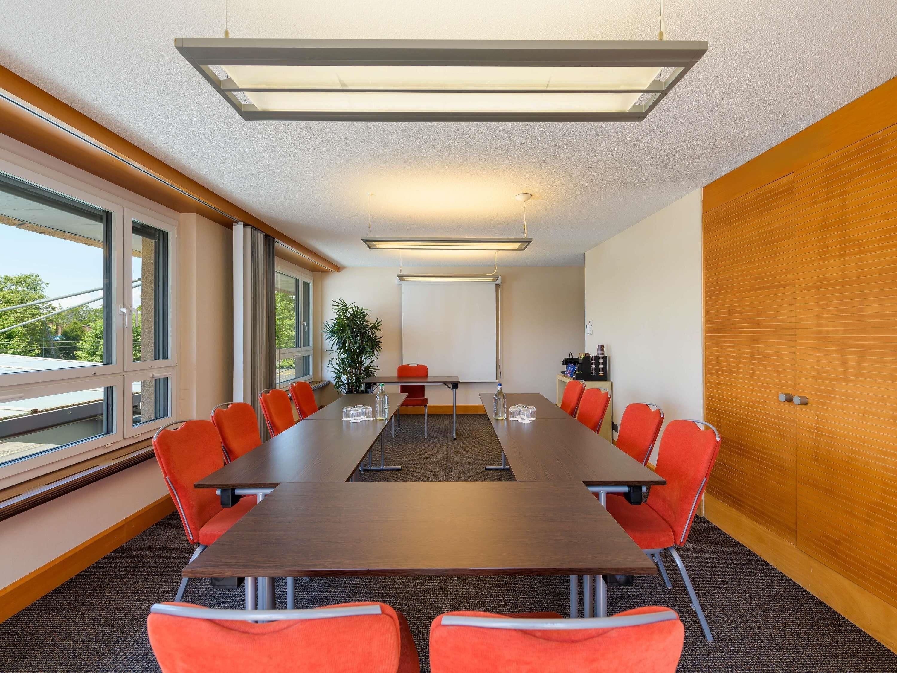 Un salón de conferencias con una larga mesa, sillas naranjas, una pantalla de proyector y ventanas amplias.