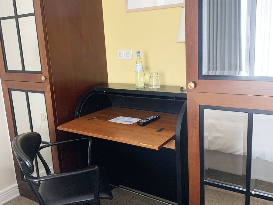 A compact room with a wooden desk, a black chair, a remote control, and a small bottle on the desk.