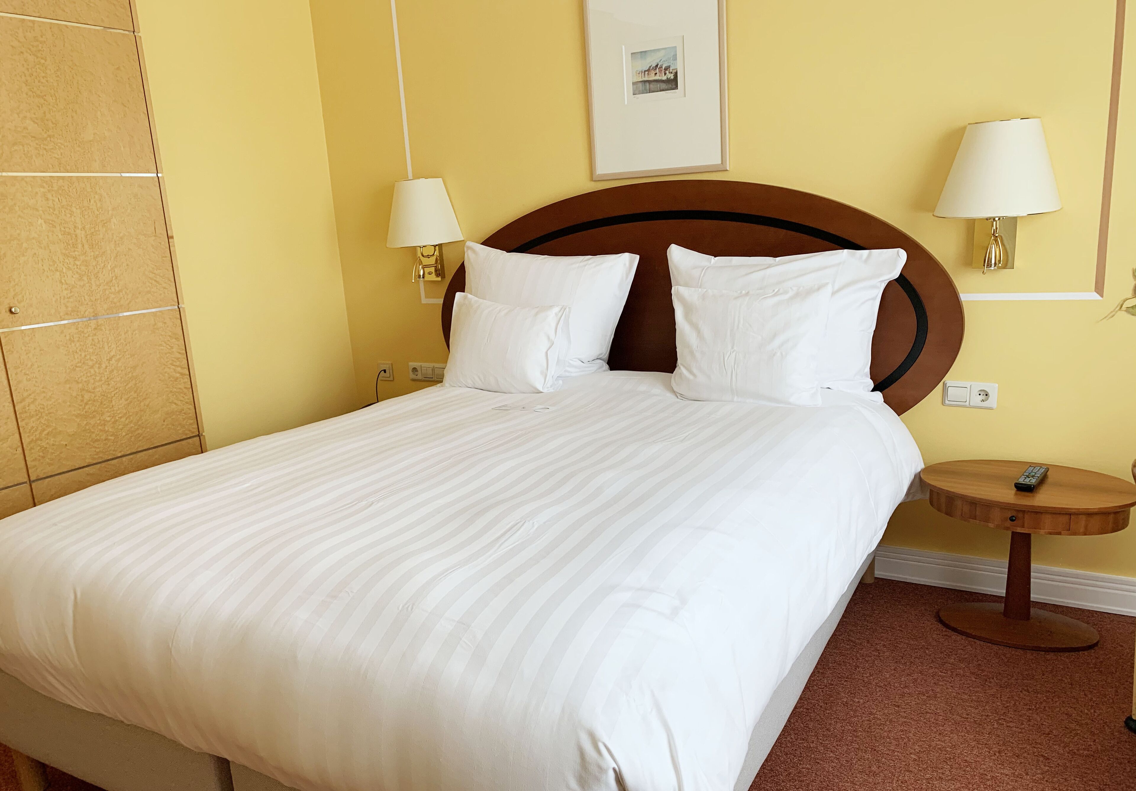 A neatly made bed with white linens, a wooden headboard, two bedside lamps, and a small wooden side table with a remote control.