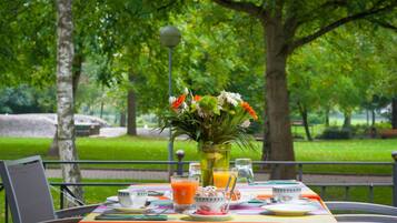 Una mesa de comedor al aire libre con un mantel colorido, un centro de mesa floral y alimentos para el desayuno, como jugo de naranja y pasteles.