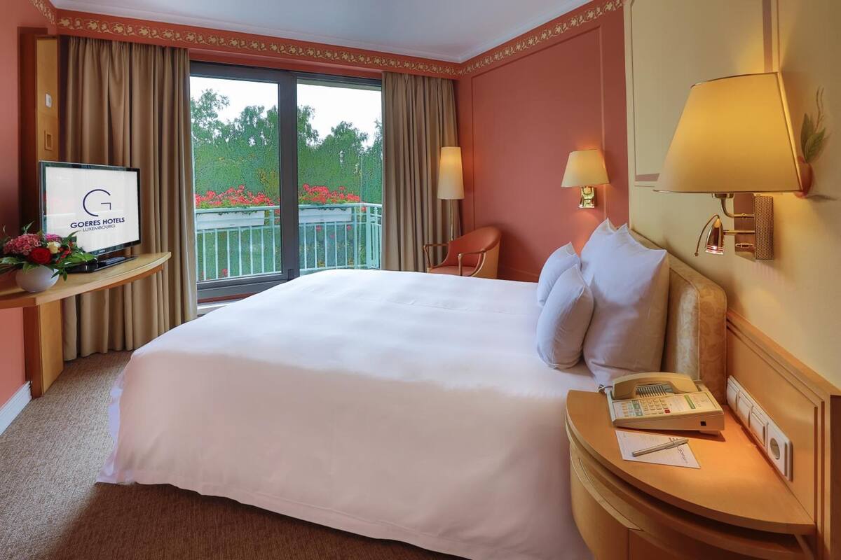 Hotelkamer met een groot bed, een televisie op een standaard, een bureau met een lamp en een balkon met uitzicht op bomen en bloemen.