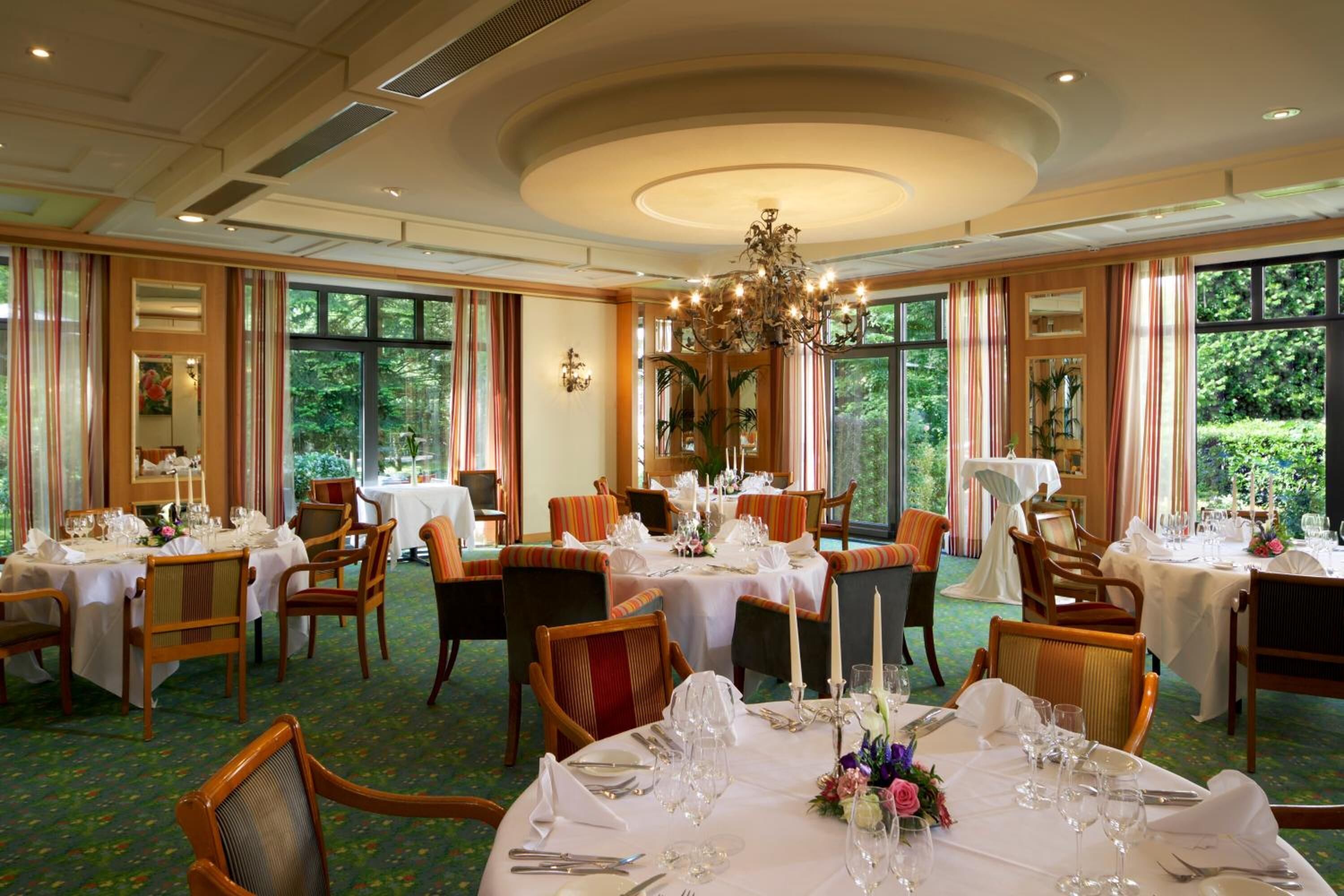 A formal dining room with multiple tables set for service, large windows with curtains, and a chandelier.