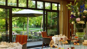 Zona de menjador amb taules preparades per al servei de te, un aparador amb diferents postres i una vista d'un jardí a través de grans finestres.