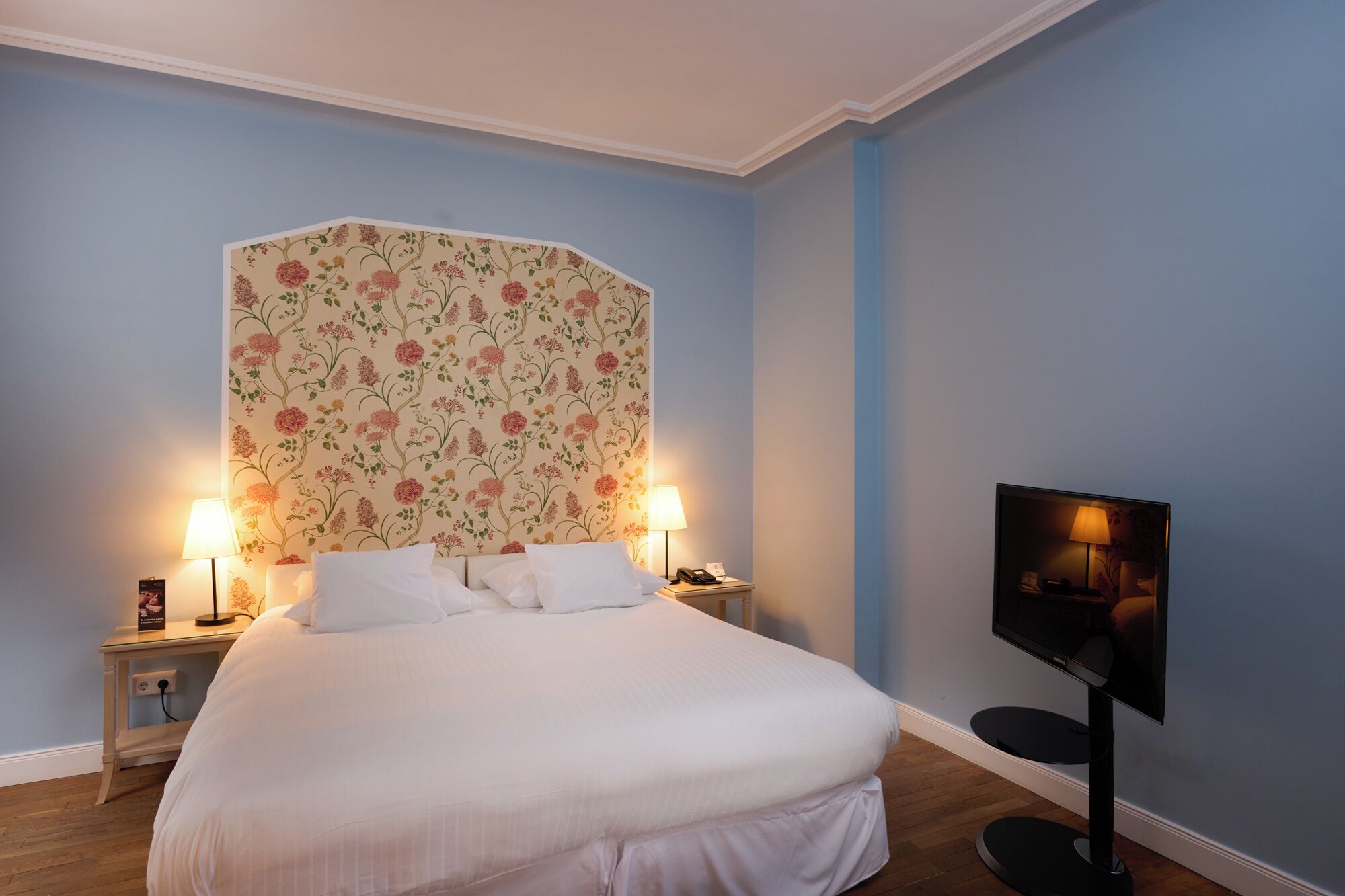 A neatly made bed with white linens, two bedside lamps, a floral-patterned headboard, and a flat-screen TV on a stand.
