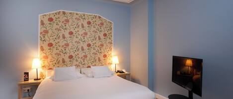 A neatly made bed with white linens, two bedside lamps, a floral-patterned headboard, and a flat-screen TV on a stand.