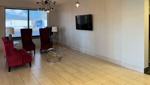 Un salon moderne avec une grande fenêtre, deux fauteuils rouges, une table en verre, un lampadaire et un téléviseur à écran plat fixé au mur.