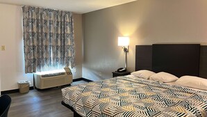 Une chambre d’hôtel avec un lit, une table de chevet, une lampe, une fenêtre avec des rideaux à motifs et un climatiseur fixé au mur.