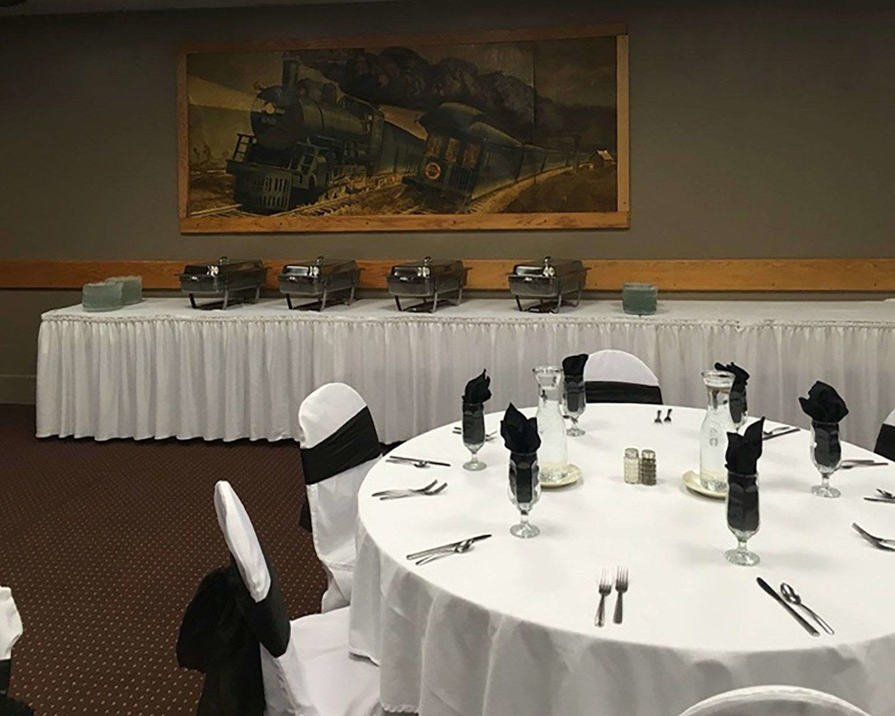 Uma sala de conferências com mesas redondas preparadas para uma reunião, toalhas brancas e capas de cadeira pretas. Um quadro grande de um trem está na parede.