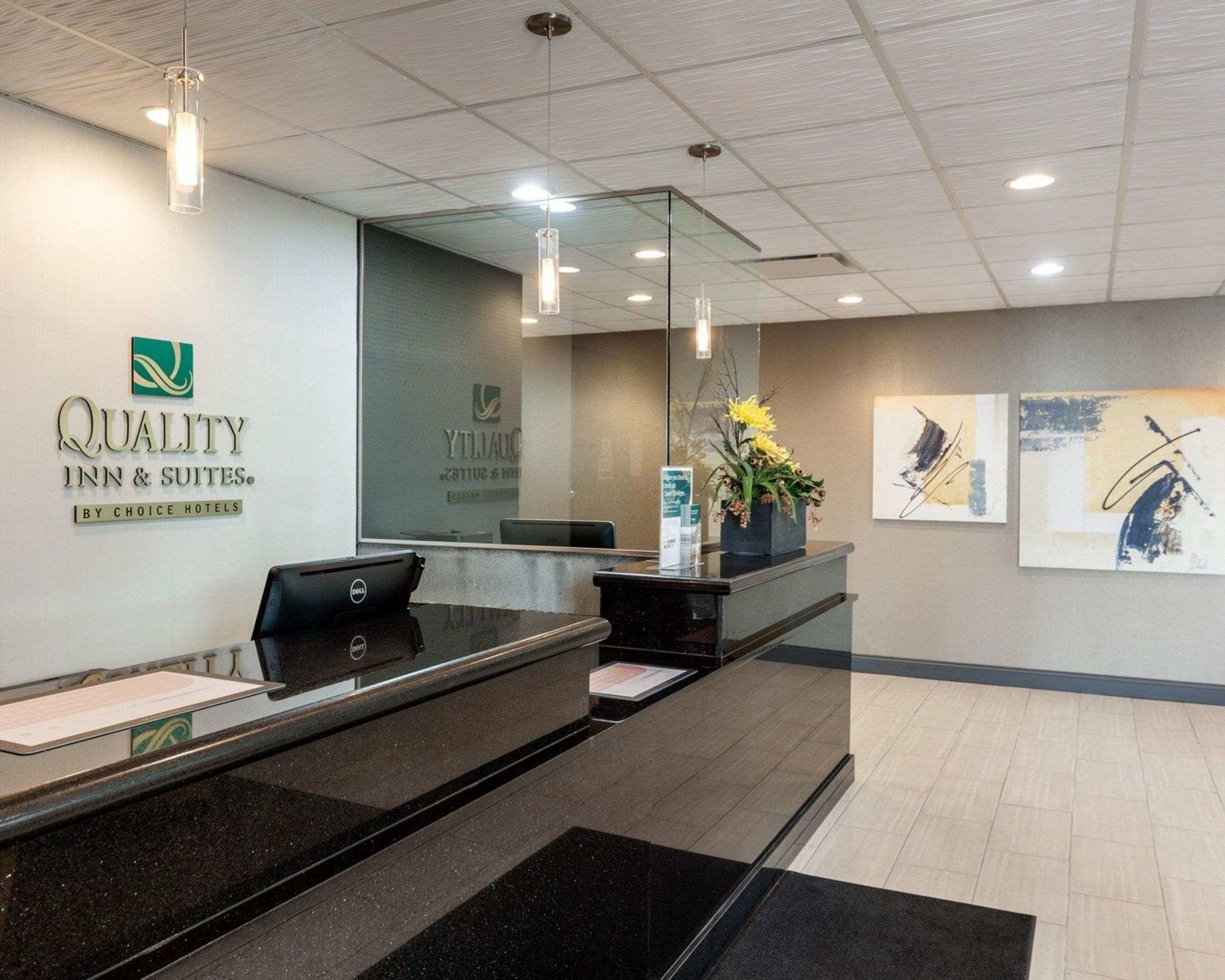 A hotel reception area with a reception desk, a computer, and a glass partition. There are framed artworks on the wall and a flower arrangement on the counter.