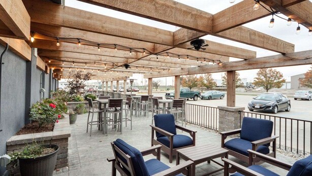 Une terrasse extérieure avec des tables et des chaises en bois, des plantes en pot et une vue sur le parking.