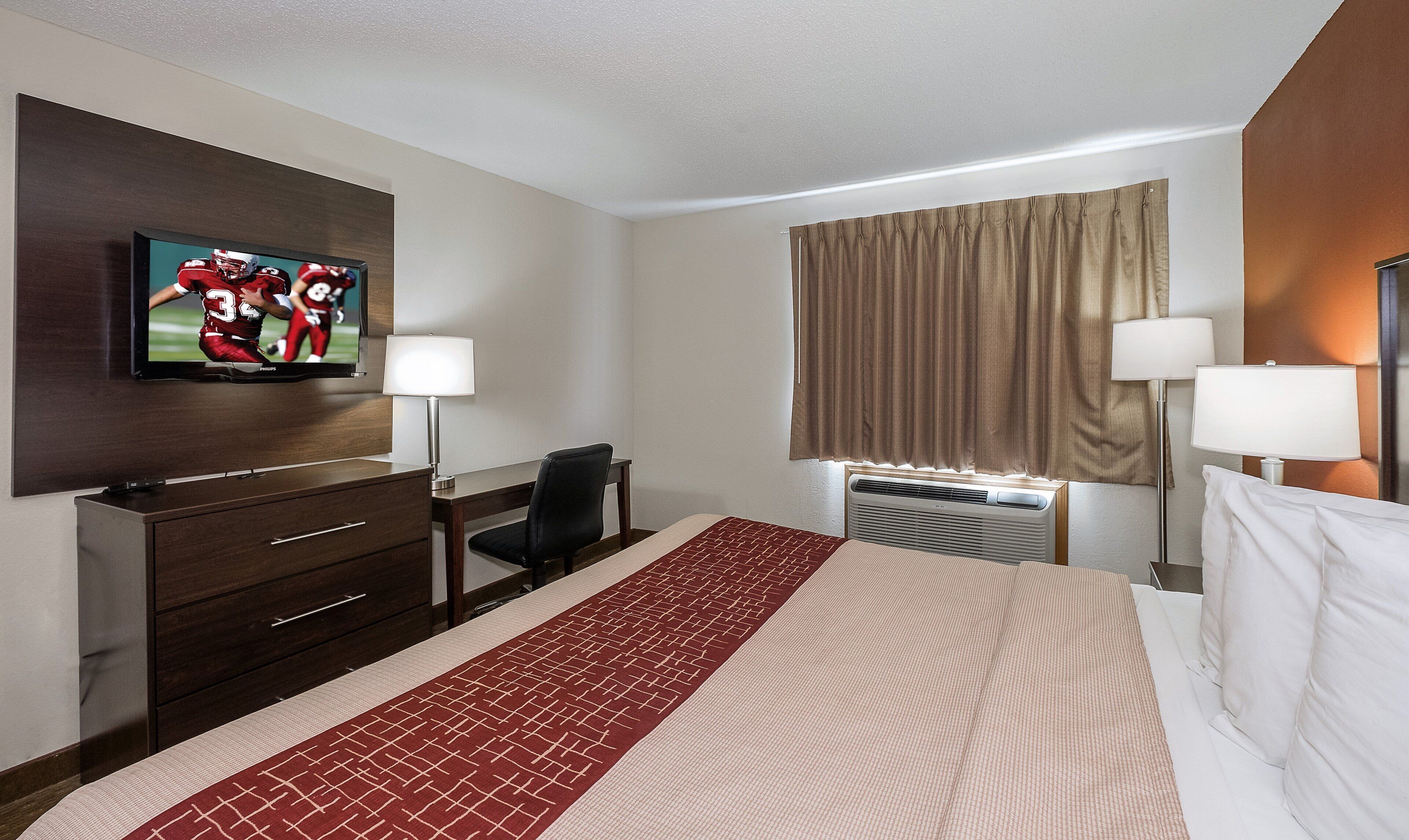 A hotel room with a bed, a desk, a chair, a television, two lamps, and an air conditioning unit.
