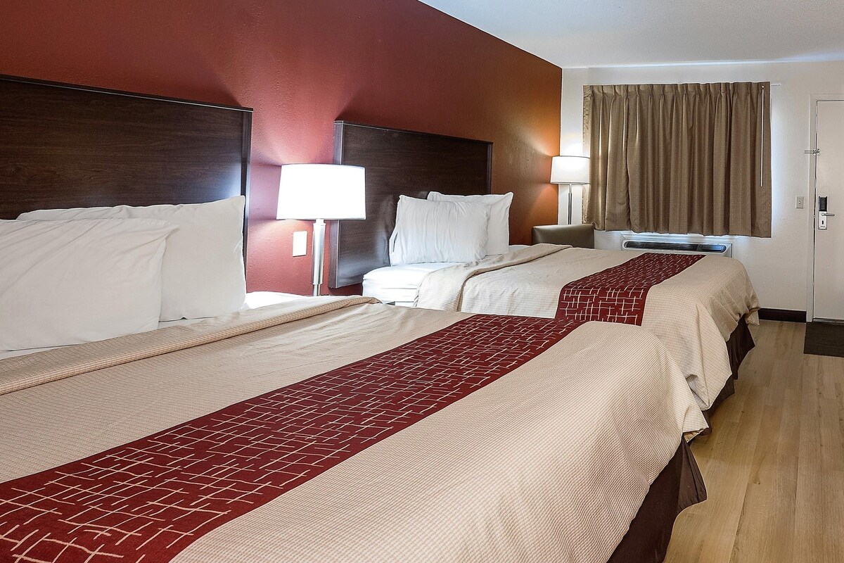 A hotel room with two beds, a nightstand, a lamp, and a window with curtains.