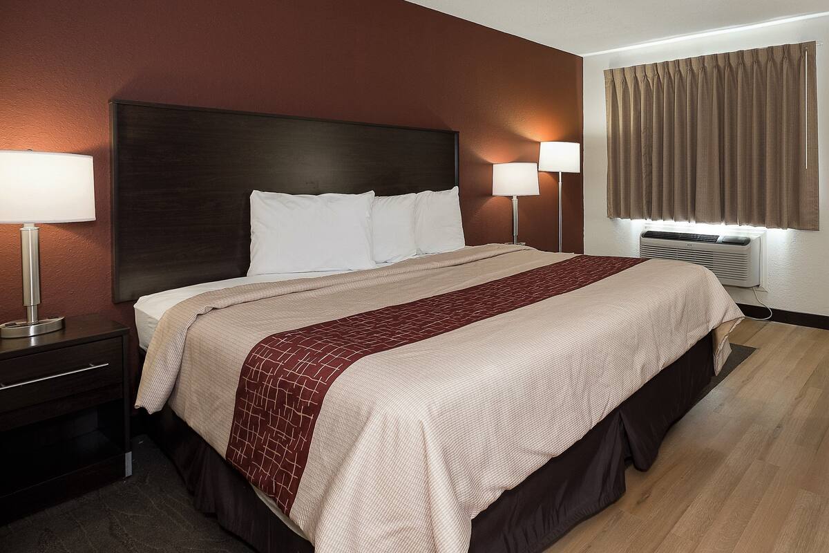 Une chambre d’hôtel avec un grand lit, deux lampes de chevet, une tête de lit sombre et une fenêtre avec des rideaux.
