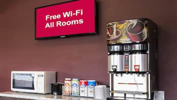 Een koffieapparaat, een magnetron en een televisie waarop staat: 'Gratis Wi-Fi in alle kamers', in een kamer met een bruine wand.