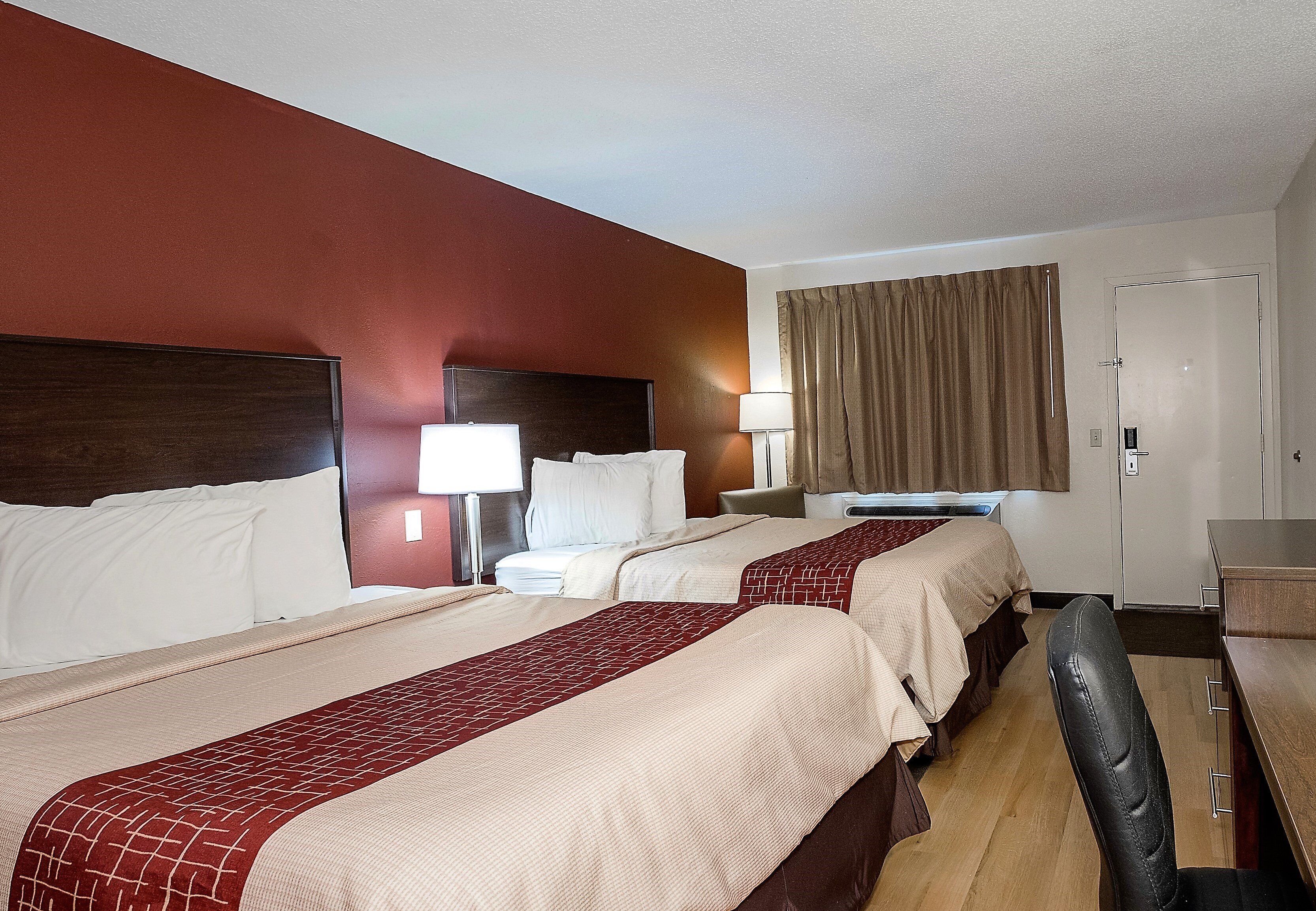 A hotel room with two beds, a desk, a chair, a window with curtains, and a door.