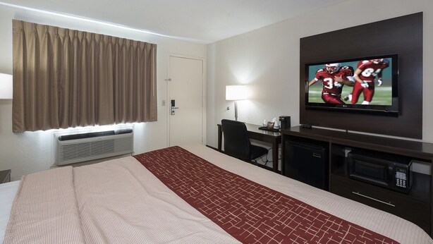A hotel room with a bed, a TV mounted on the wall, a desk with a lamp, and a minibar.