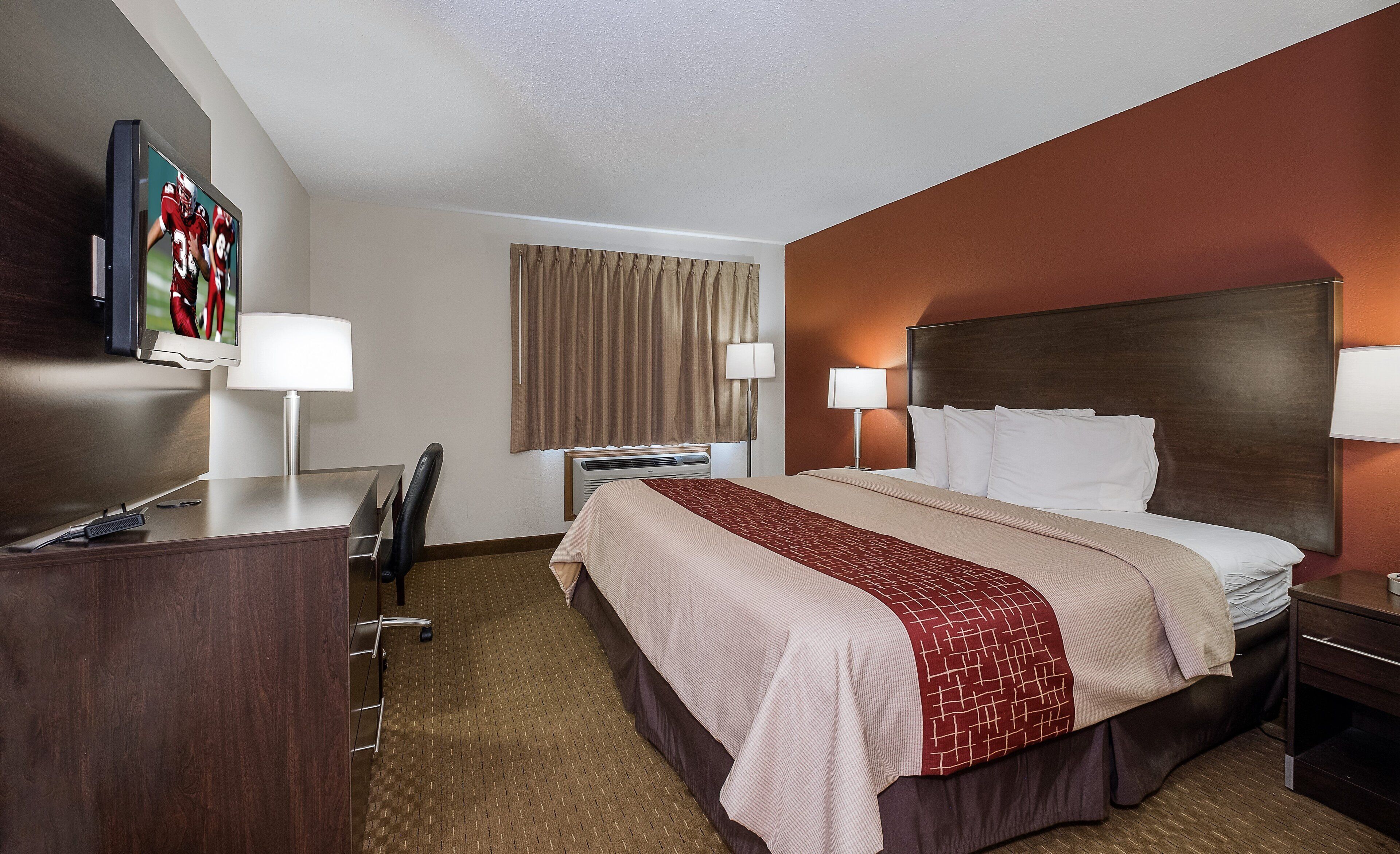 A hotel room with a bed, bedside lamps, a desk with a chair, a television, and a window with curtains.