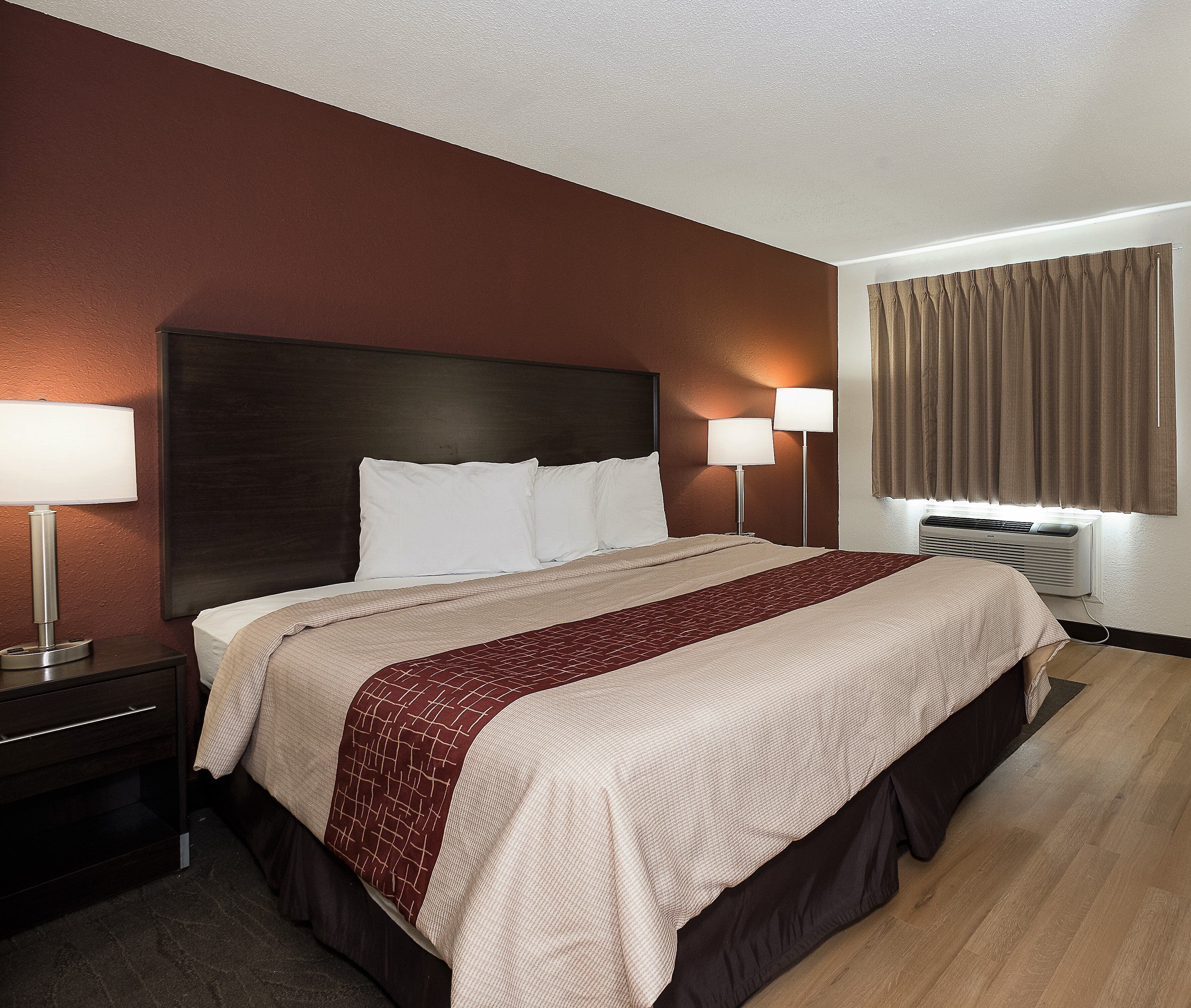 Quarto de hotel com uma cama grande, duas luminárias laterais, cabeceira escura, janela com cortinas e ar-condicionado fixado na parede.