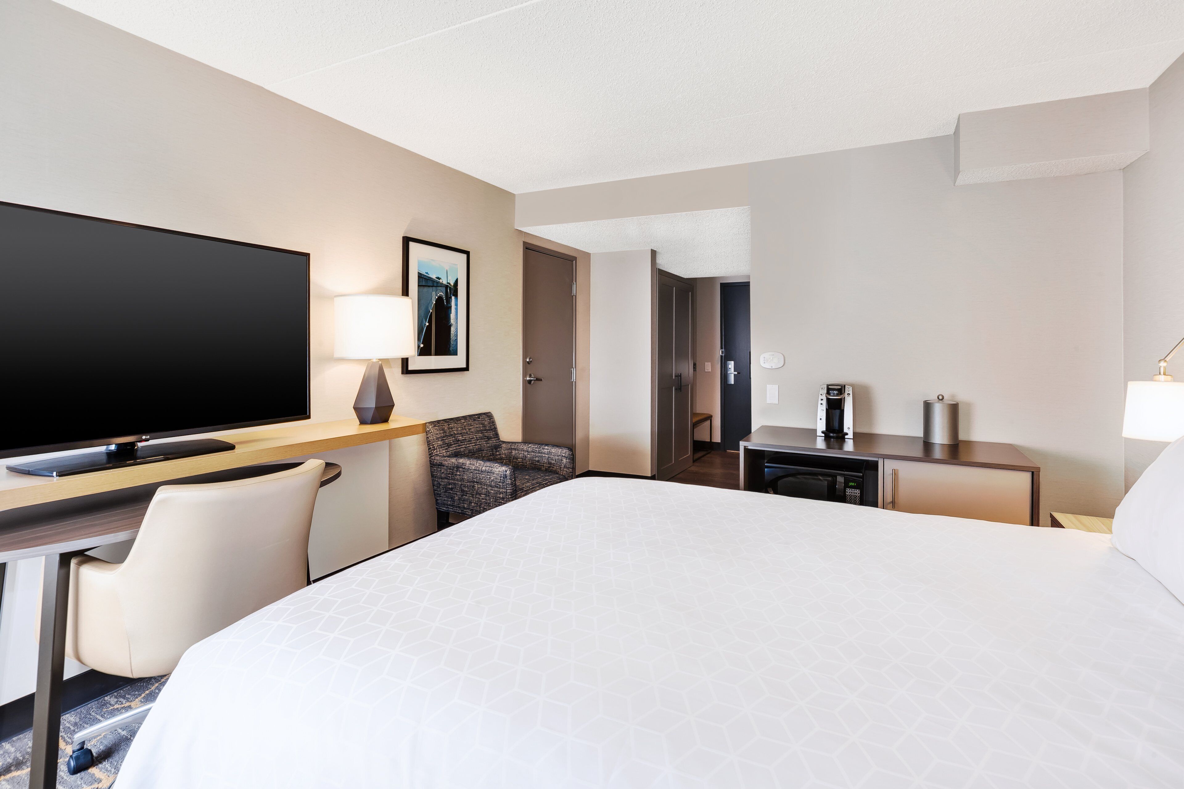 A hotel room with a large bed, a desk with a flat-screen TV, a chair, a coffee maker, and a lamp.