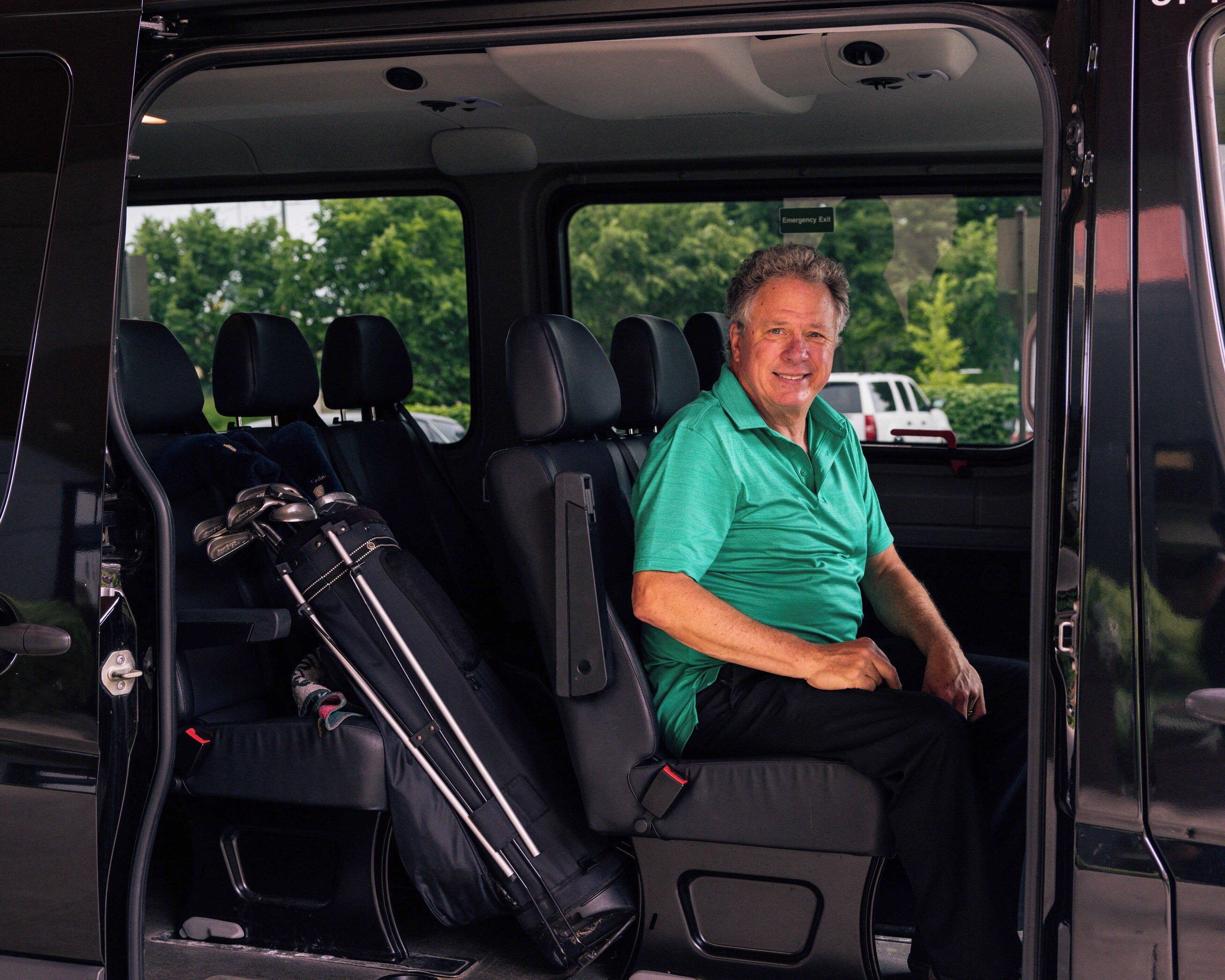 A man seated in a van with luggage and golf clubs in the back.