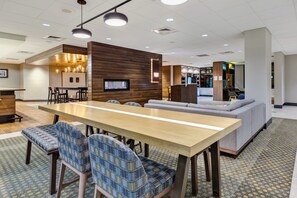 A modern hotel lobby with a long communal table, seating area, and a bar.