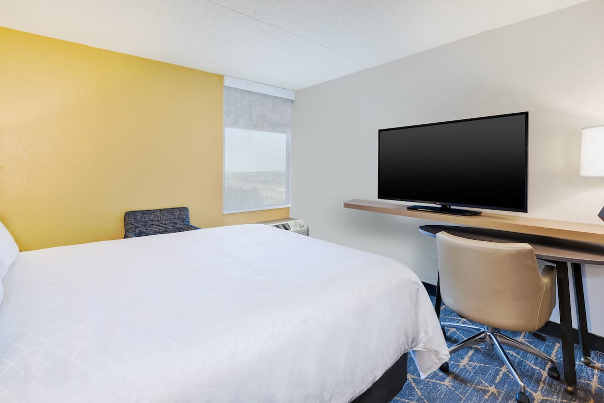 A hotel room with a large bed, a desk with a flat-screen TV, a chair, and a window with blinds.
