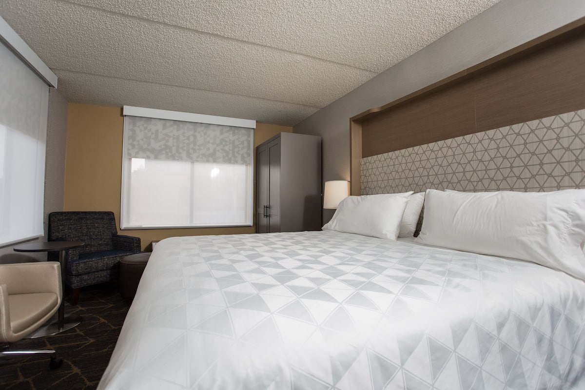 A hotel room with a large bed, a desk, a chair, a wardrobe, and a window with blinds.