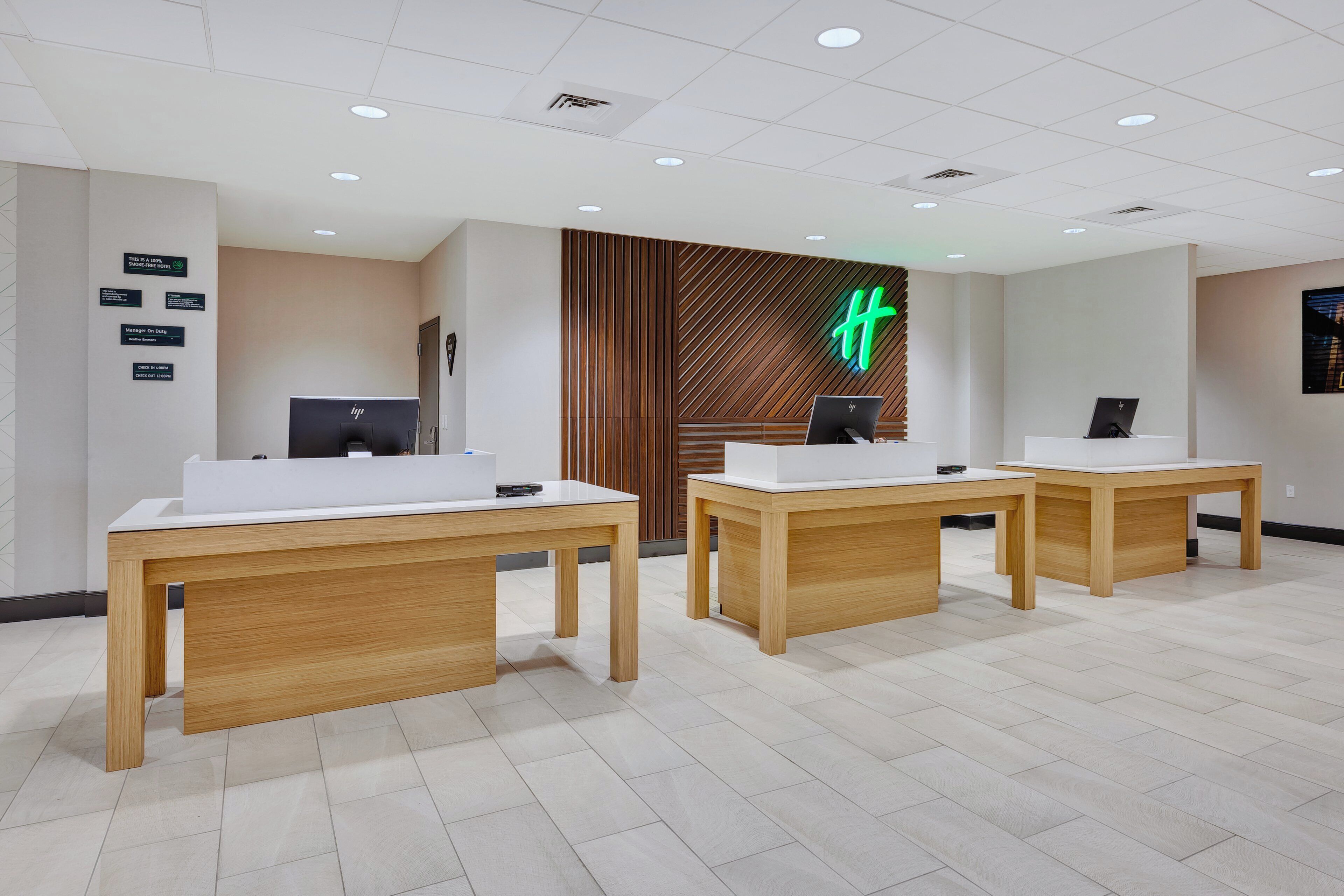 A hotel reception area with two reception desks, a wooden panel wall with a neon sign, and a ceiling with recessed lighting.