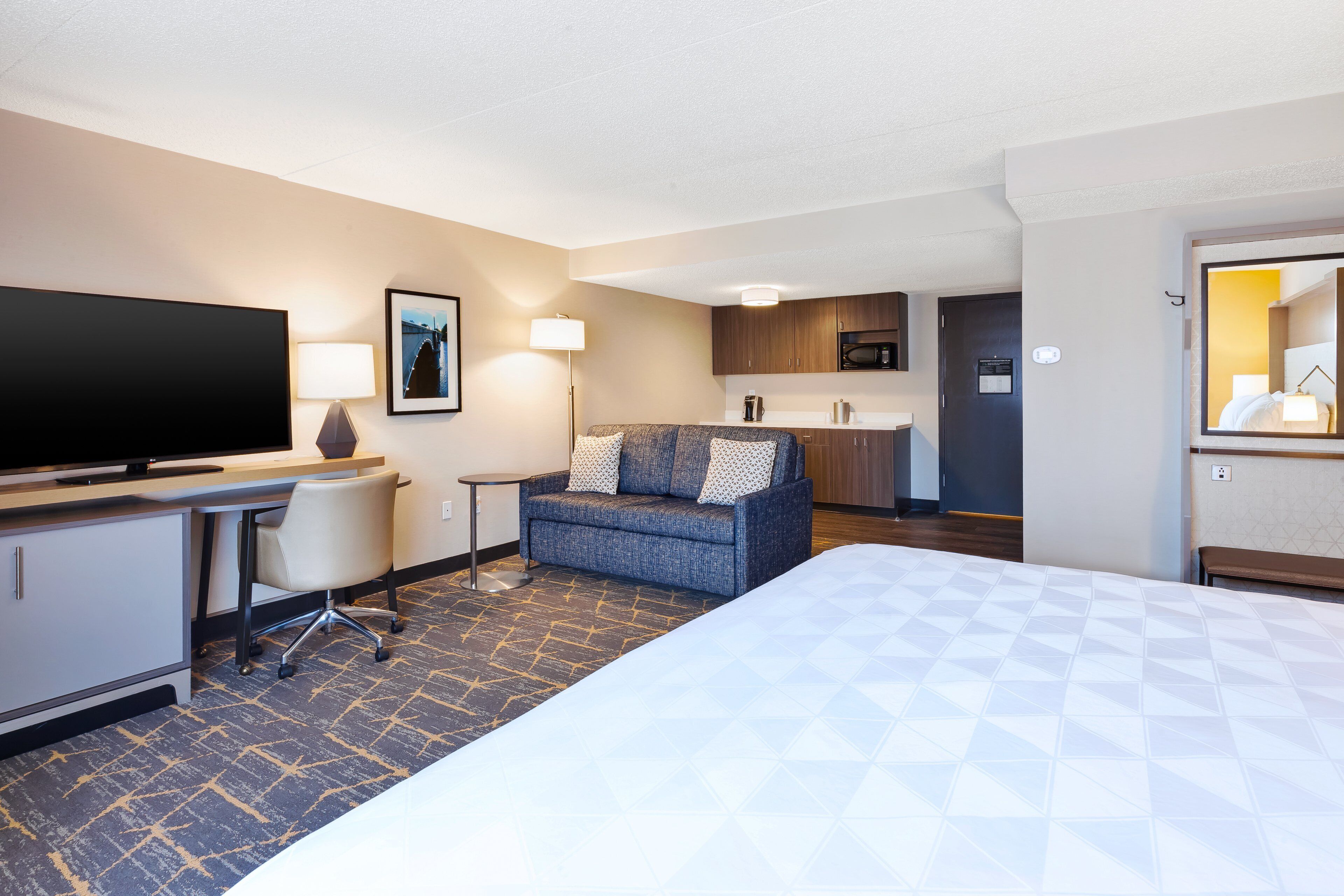 A hotel room with a bed, a desk, a chair, a sofa, a TV, a lamp, a microwave, and a small kitchen area.