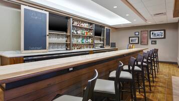 A bar area with a long counter, multiple shelves of bottles, glasses, and a television. There are several chairs around the bar.