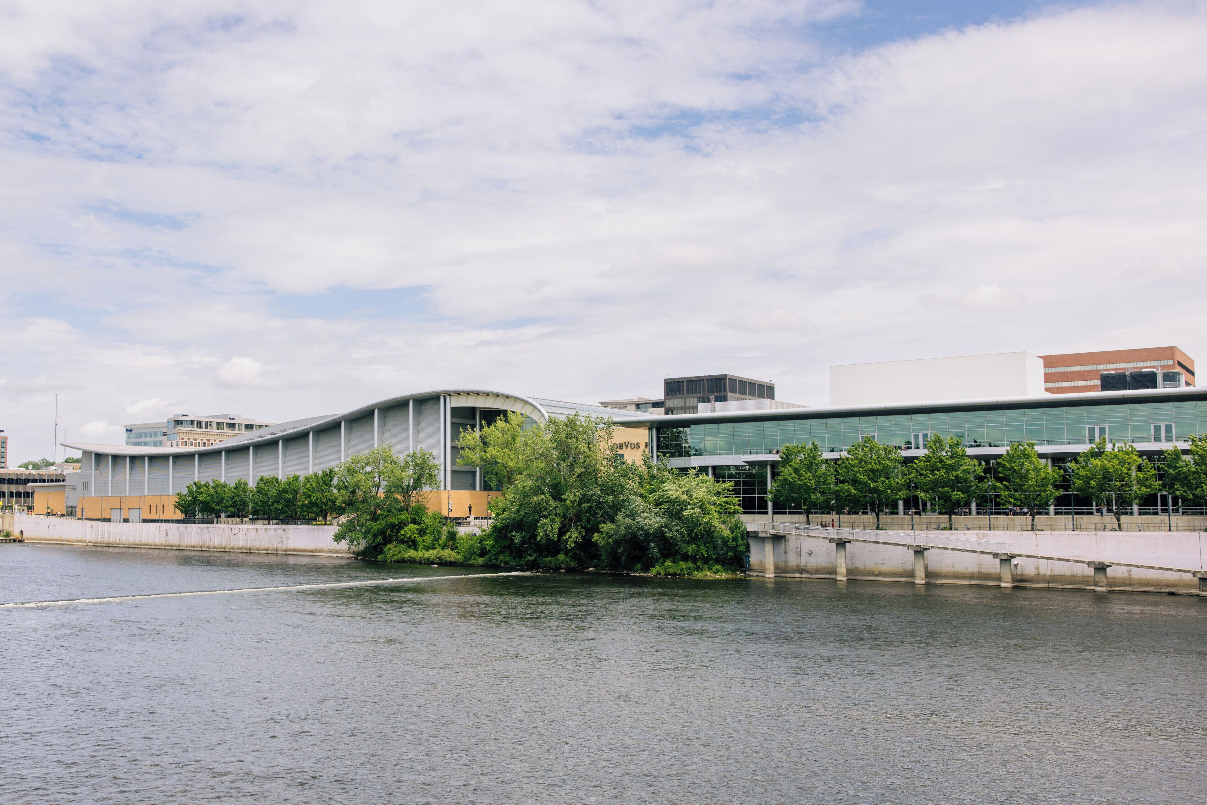 A modern building with a curved facade, a river in the foreground, and a bridge connecting to another structure.