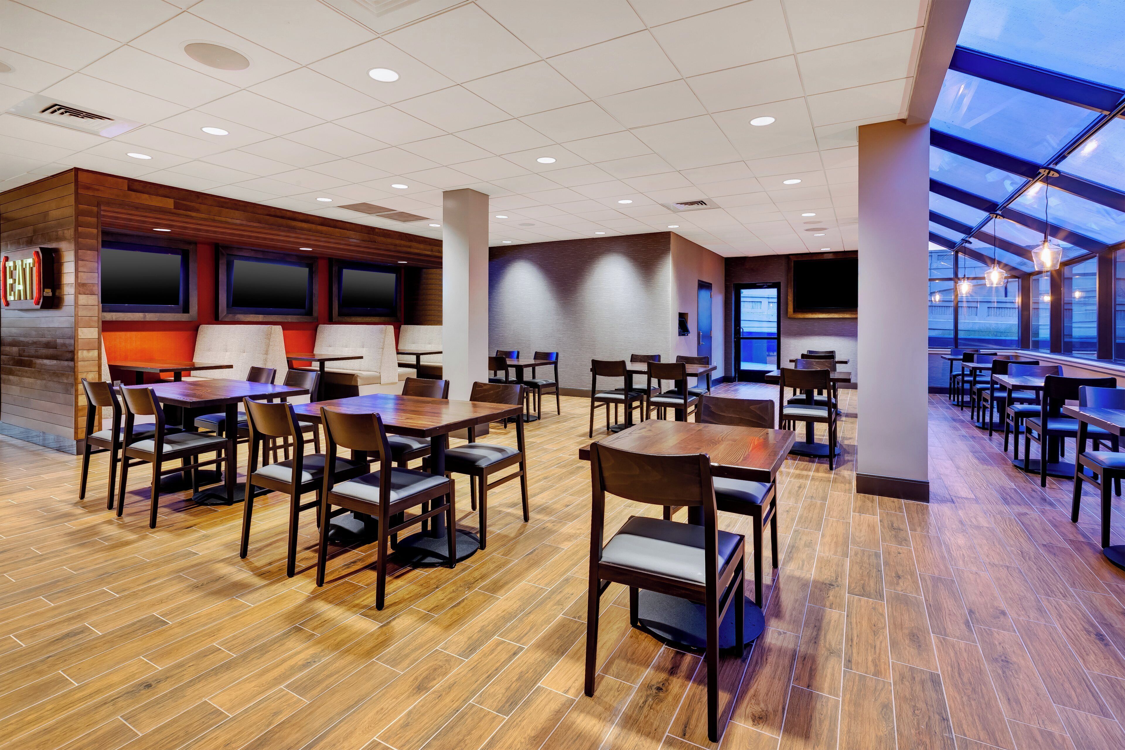 A spacious restaurant with multiple dining tables and chairs, a wooden floor, and a large window overlooking the city.