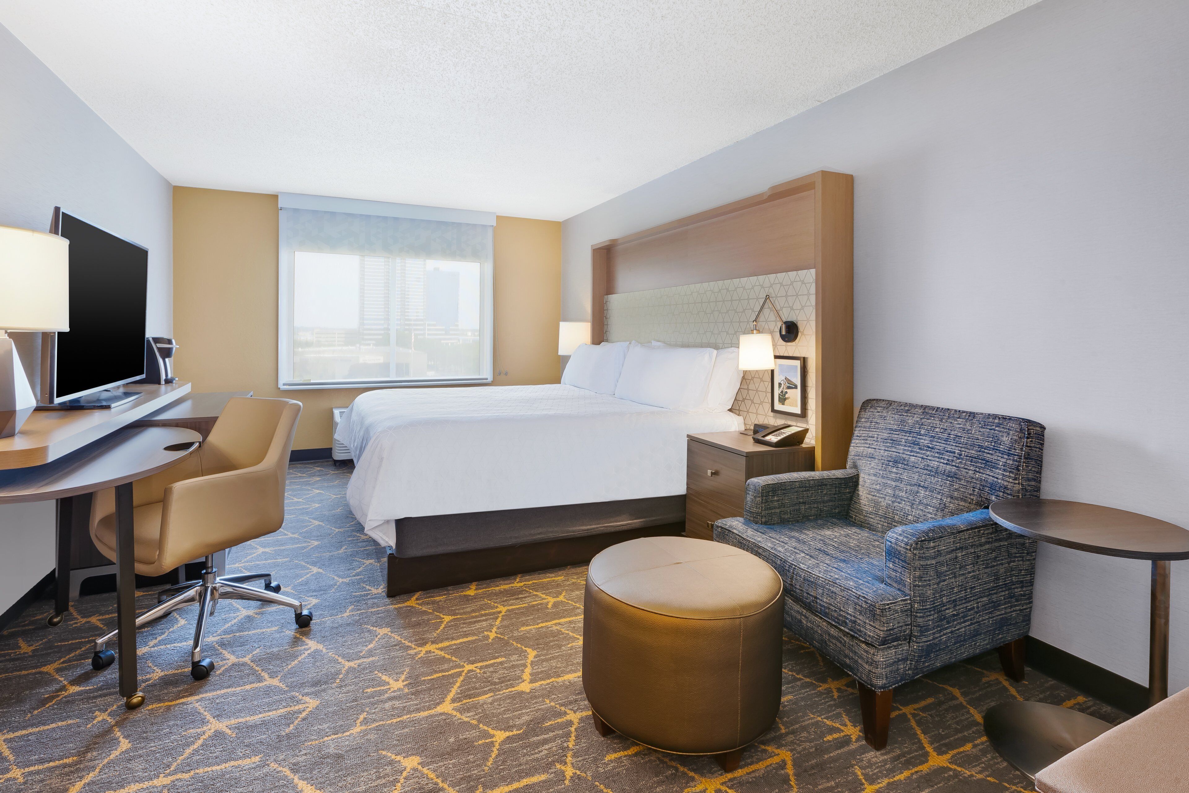 A hotel room with a bed, a desk with a chair, a round ottoman, a small round table, and a blue armchair.