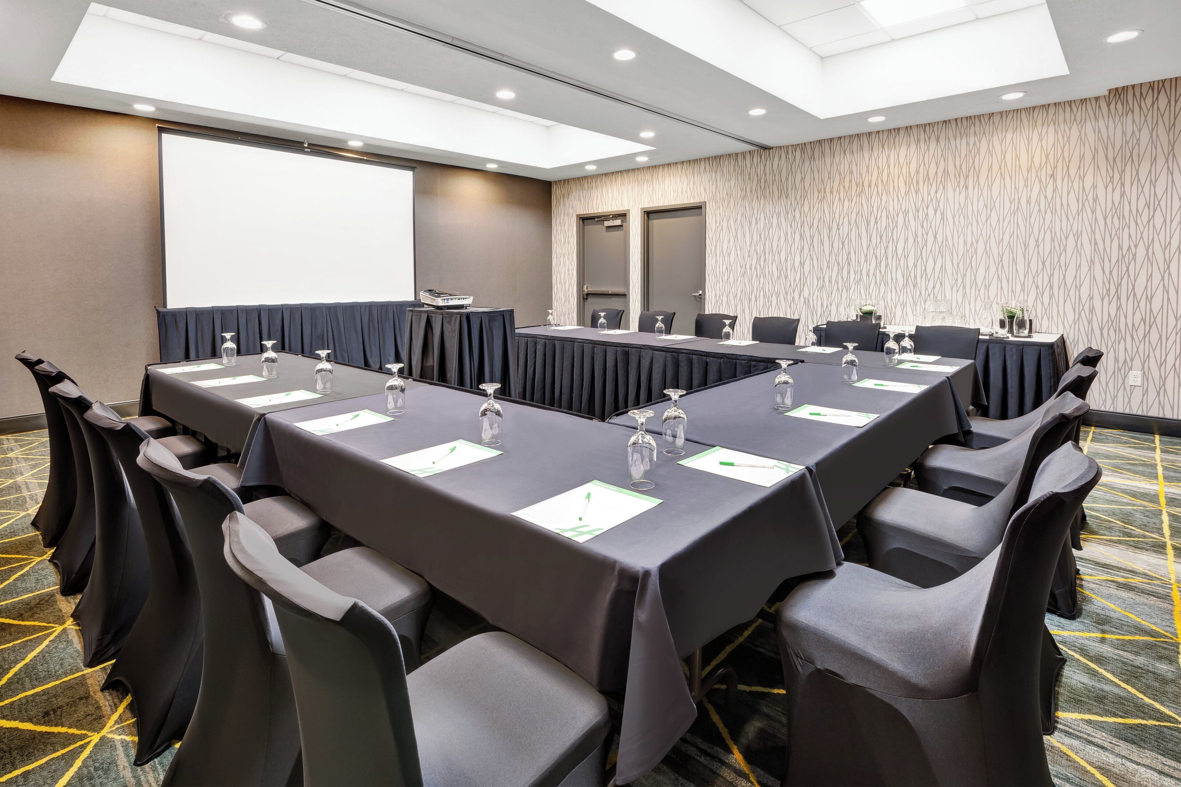 Een vergaderzaal met een lange tafel, stoelen, een scherm voor een projecter en een kamerbreed tapijt met een patroon.