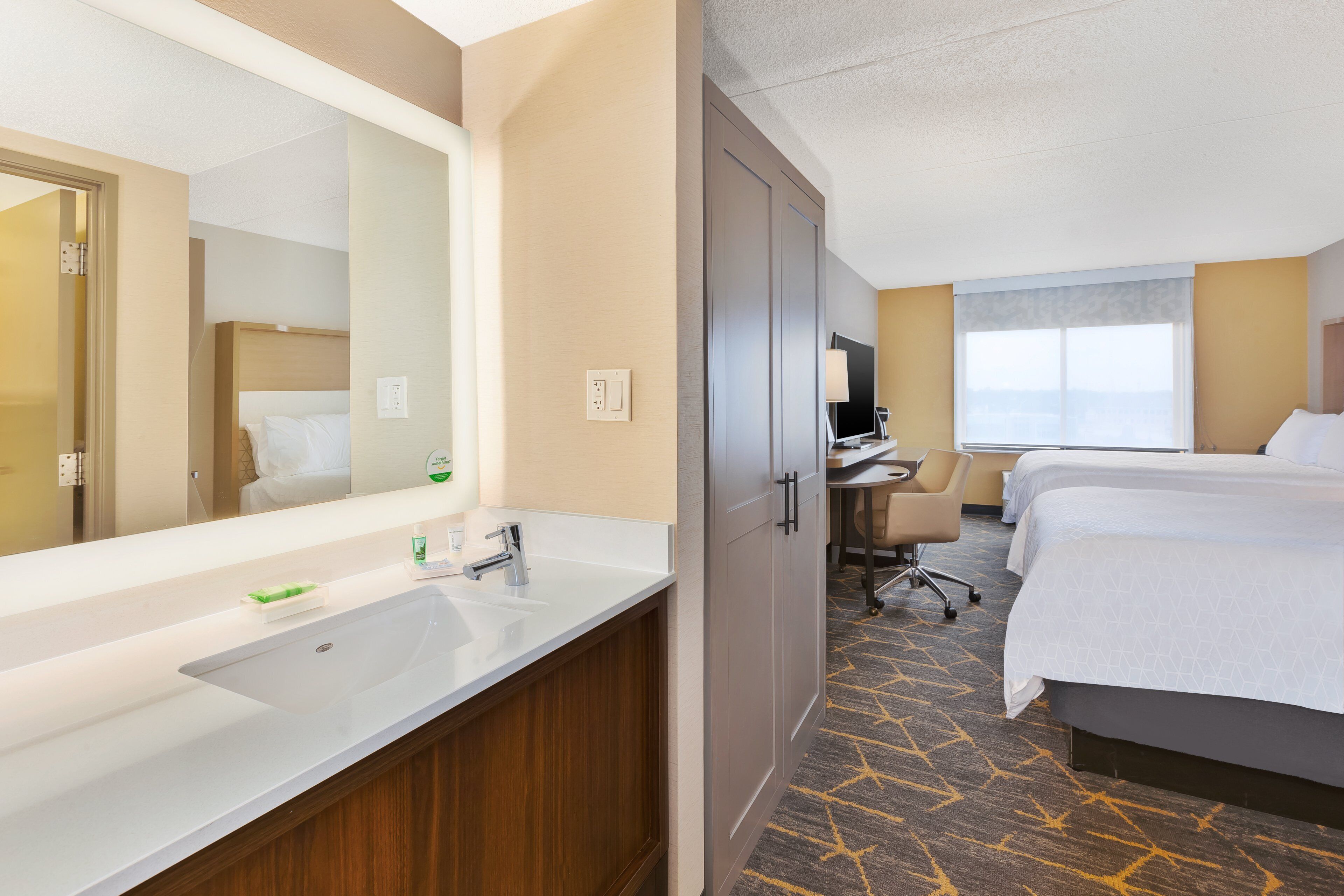 Quarto de hotel com um espelho grande, pia, secretária, duas camas e janela com vista.