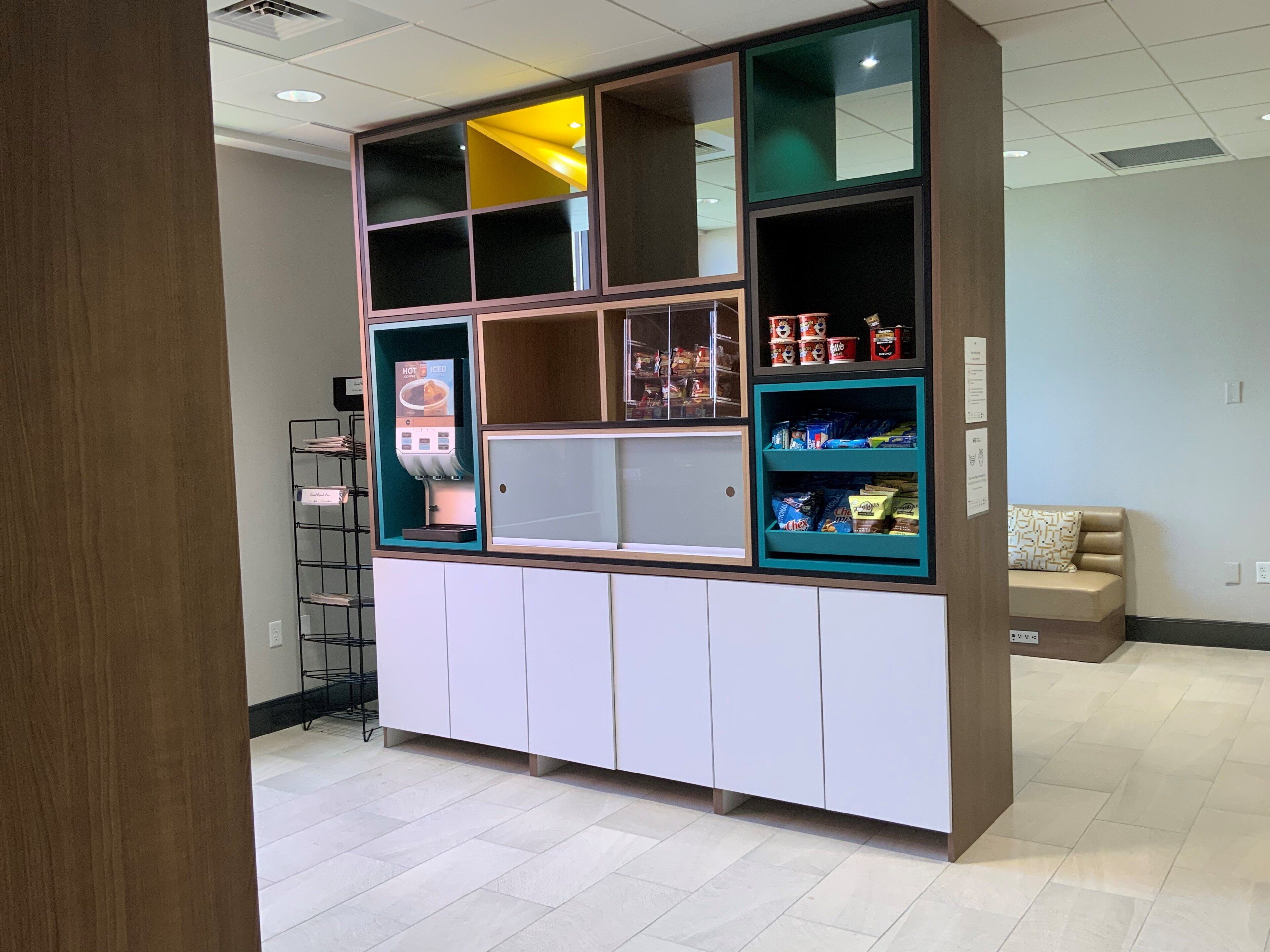 A kitchenette with a coffee machine, shelves with snacks, and a seating area with a patterned cushion.