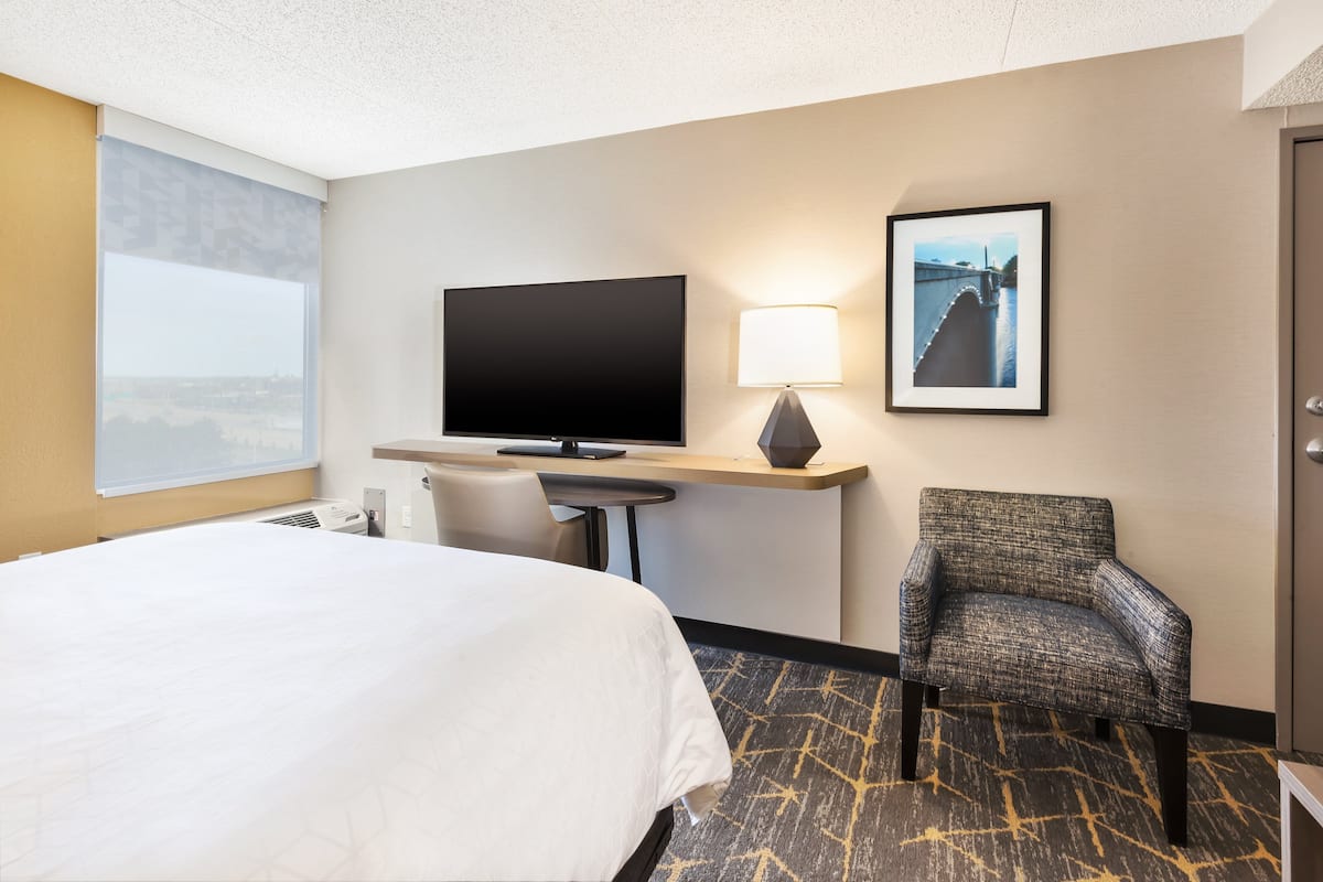 A hotel room with a large bed, a flat-screen TV, a desk with a lamp, a framed picture on the wall, and a chair.