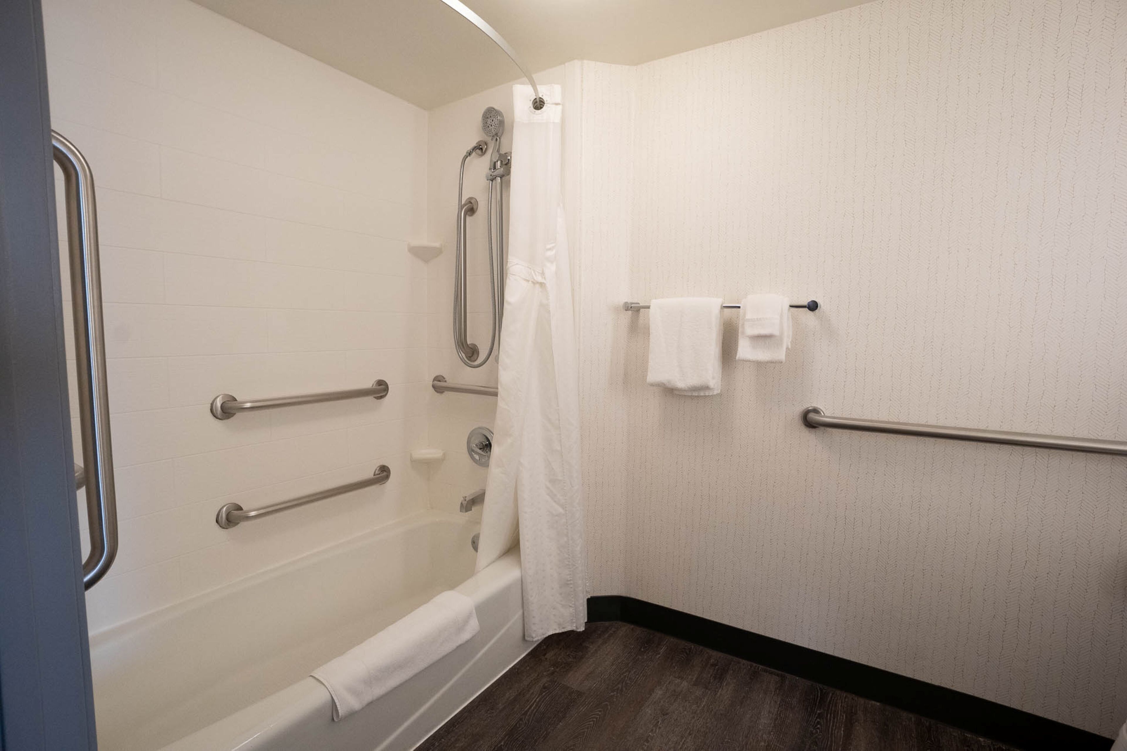 A bathroom with a shower, grab bars, a towel rack, and a wooden floor.