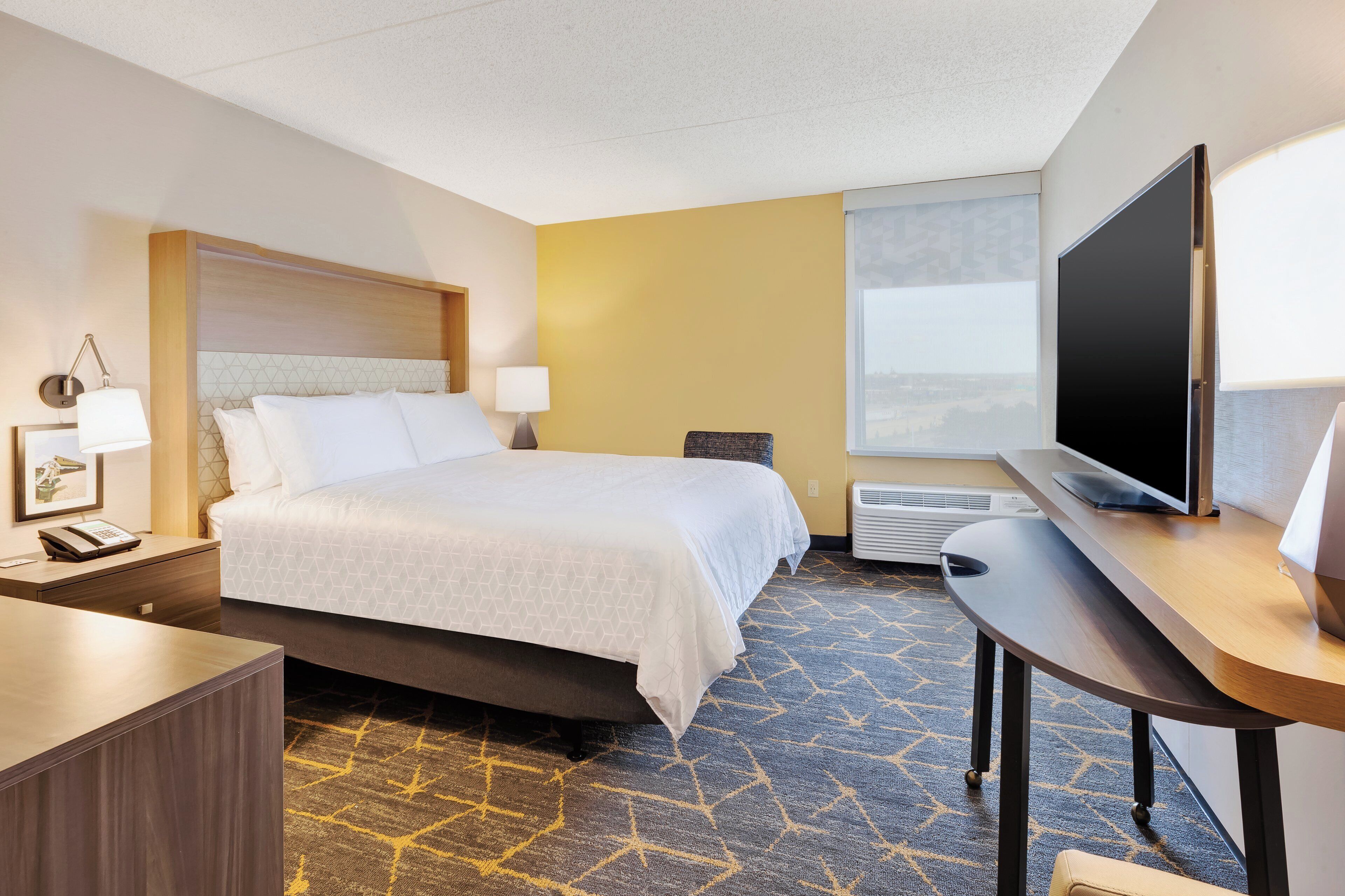 A hotel room with a bed, bedside table, lamp, TV, and a view of the city.