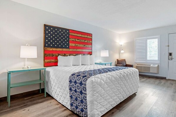A hotel room with a bed, two bedside tables, a wall-mounted American flag, a window with blinds, and an air conditioning unit. - Rodeway Inn & Suites Fort Rucker (Daleville)