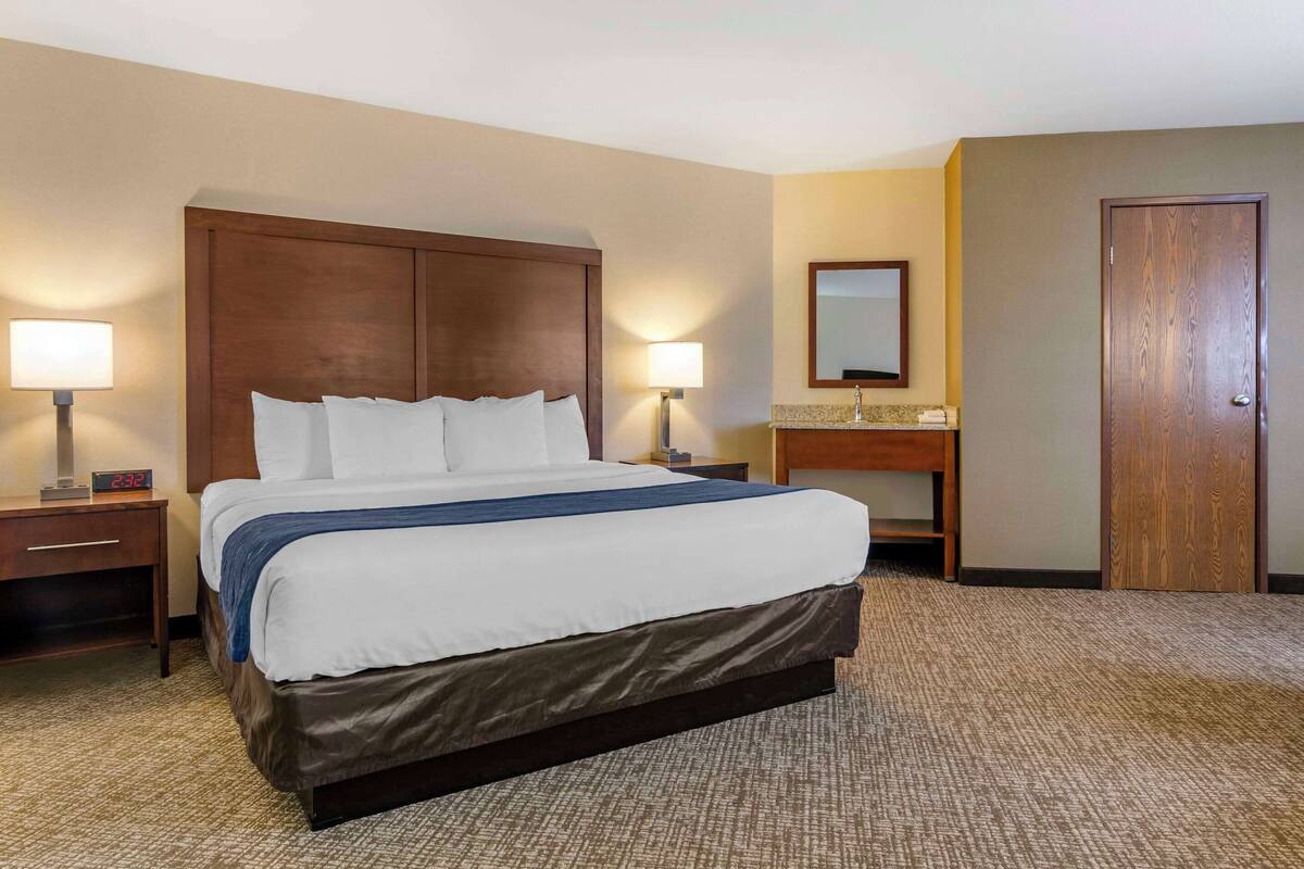 A hotel room with a large bed, two bedside tables with lamps, a wooden headboard, a bathroom with a sink and mirror, and a door.
